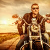 Biker man wearing a leather jacket and sunglasses sitting on his motorcycle looking at the sunset.